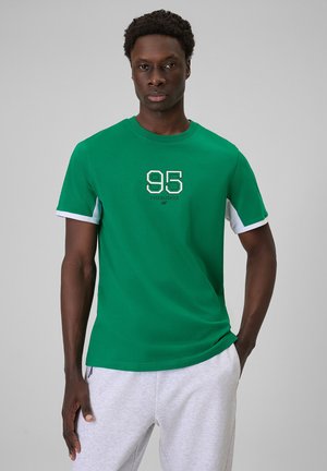 Mężczyzna ubrany w zielony T-shirt z białymi akcentami na rękawach i napisem „95 Established”, w połączeniu z jasnoszarymi spodniami dresowymi, stoi na szarym tle.