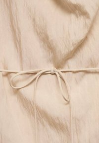 Tessuto beige con una leggera texture e pieghe sottili, caratterizzato da un dettaglio di cintura annodata dello stesso colore. Messa a fuoco sul materiale liscio e leggero.