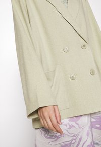 Světle zelený texturovaný dvouřadý blazer s velkými knoflíky a širokými rukávy, doplněný vzorovaným levandulovým spodním oděvem.