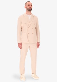 Giacca di abito beige doppiopetto con due tasche frontali e pantaloni abbinati. Indossata sopra una maglietta bianca, abbinata a sneaker chiare.