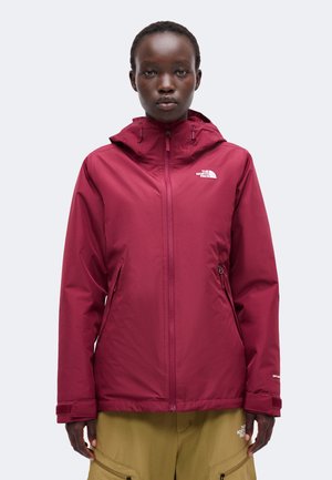 Osoba ubrana w bordową kurtkę z kapturem North Face z zamkiem z przodu i spodnie khaki na jednolitym jasnym tle.