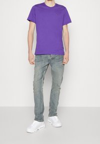 Man i en enkel lila t-shirt med kort ärm, urblekta blå jeans och vita sneakers mot en helt vit bakgrund.