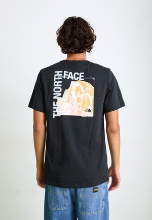 Osoba z kręconymi włosami nosząca czarną koszulkę The North Face z grafiką góry i logo na plecach, w połączeniu z luźnymi niebieskimi dżinsami.