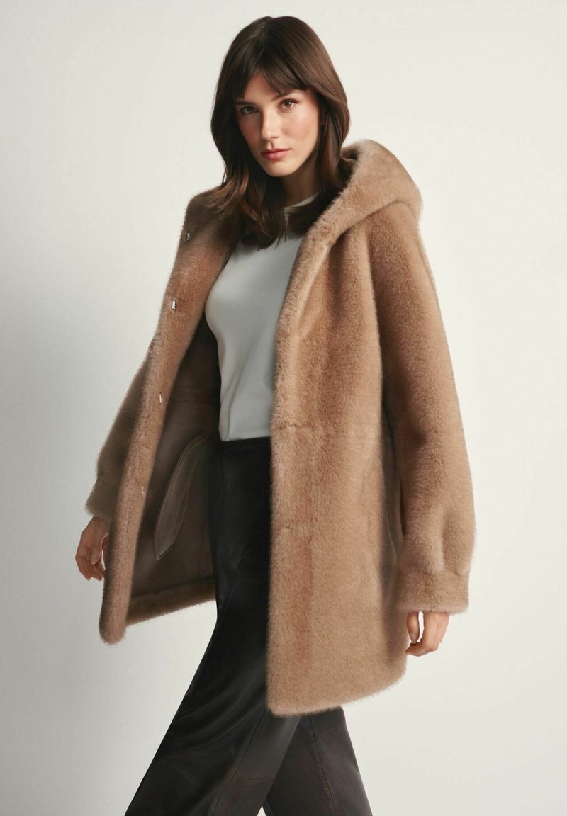 Kobieta ubrana w kremowy puchaty płaszcz z kapturem, na białej bluzce i czarnych spodniach, stojąca na neutralnym jasnym tle.