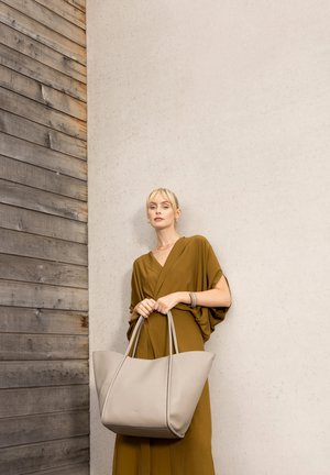Femme en robe moutarde tenant un grand sac fourre-tout en cuir beige, debout devant un mur en bois et béton.