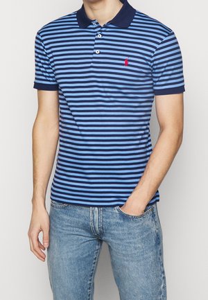Mann trägt blau- und marineblau gestreiftes Poloshirt mit rotem Logo und hellblauer Jeans, rechte Hand in der Tasche, steht vor neutralem Hintergrund.