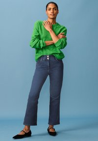 Roheline kudumisel sweater, millel on paks tekstuur, on kombineeritud tumehallide laienevate teksadega, millel on kaks kuldset nuppu. Mustad lamedad jalanõud täiendavad välimust.