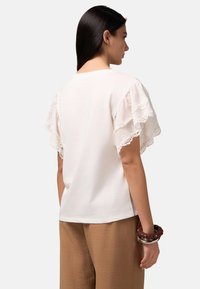 T-shirt bianco con maniche in pizzo a volant, caratterizzato da una vestibilità rilassata e scollatura arrotondata. Indossato con pantaloni marrone chiaro texturizzati e braccialetti impilati.