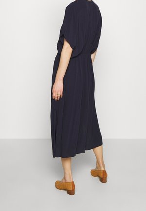 Robe midi bleu marine à coupe fluide, taille élastique et manches courtes. Associée à des chaussures à talons blocs tressées beiges. Tissu au toucher lisse.