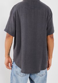 Camicia a maniche corte in grigio scuro, caratterizzata da un motivo testurizzato e una vestibilità rilassata. L'orlo è leggermente curvo con spacchetti laterali per facilitare il movimento.