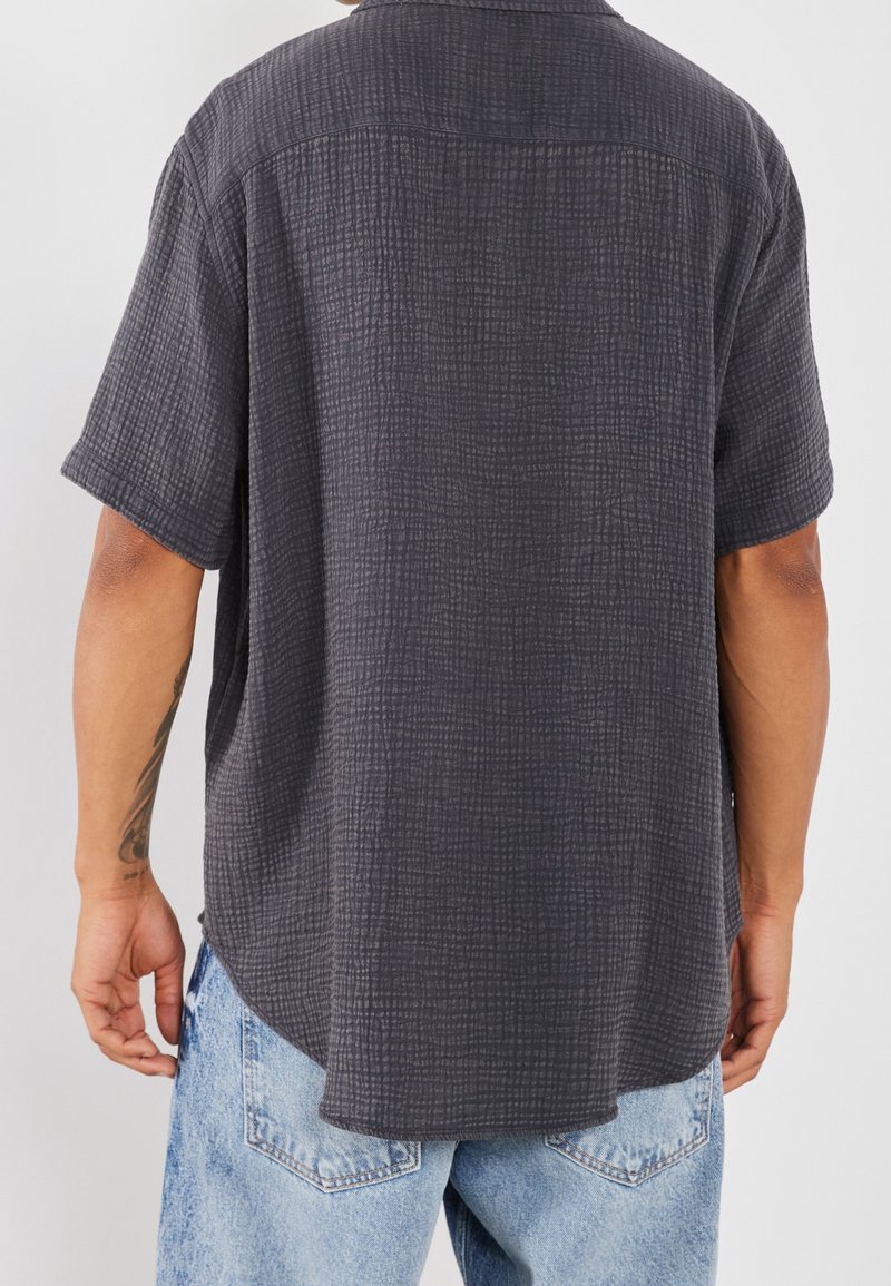 Camicia a maniche corte in grigio scuro, caratterizzata da un motivo testurizzato e una vestibilità rilassata. L'orlo è leggermente curvo con spacchetti laterali per facilitare il movimento.