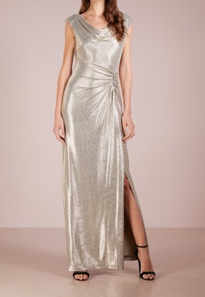 Srebrna metaliczna suknia bez rękawów z marszczonym dekoltem, marszczeniem po bokach z ozdobami, rozcięciem do uda, noszona z czarnymi szpilkami na kostkę.