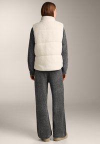 Weißer, flauschiger Weste mit hohem Kragen, kombiniert mit einem grauen, strukturierten Outfit. Der Look wird durch beigen Slip-On-Schuhwerk vervollständigt.