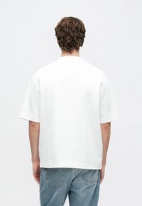 Ungdom med kort krøllete brunt hår iført en ensfarget hvit kortermet skjorte og lyseblå jeans, sett fra bakgrunnen på en hvit bakgrunn.