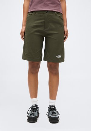 Person som har på seg olivengrønne The North Face-shorts, lilla topp, hvite sokker og svarte og grå tursko, stående mot en ensfarget bakgrunn.