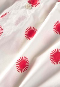 Napillispaita valkoisesta kankaasta, jossa on punaisia auringon säteiden kuvioita hajallaan. Pehmeä pinta, kaartuva kaulus ja edessä napillinen suljettava yksityiskohta.