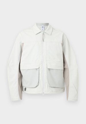 Svetlo siva jakna z zadrgo spredaj, dvema velikima sprednjima žepoma s poklopci, bež stranskima paneloma in ovratnikom v stilu srajce.