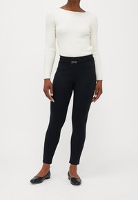 Svarta, stretchiga leggings med en slät yta och en åtsittande design, försedda med ett smalt bälte med en dekorativ metallspänne. Bärs med en vit ribbad topp.