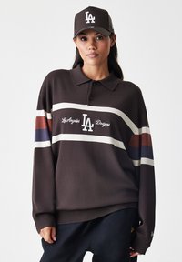 Bruine lange mouwen polo met een kraag, voorzien van witte en gekleurde strepen, en geborduurd 'LA' logo. Gecombineerd met een bijpassende bruine pet.