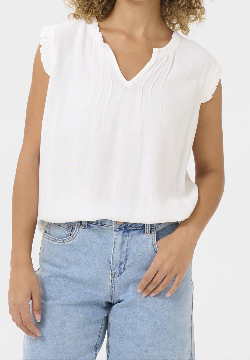 Femme portant un chemisier blanc sans manches avec des bords à volants et un jean taille haute bleu clair, main posée négligemment dans la poche.