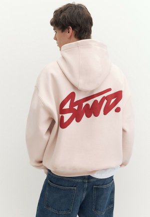 Jeune homme portant un sweat à capuche rose clair avec un grand motif graphique rouge et un jean bleu, vu de dos sur un fond uni.