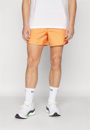 Mężczyzna stoi ubrany w jaskrawo pomarańczowe sportowe spodenki, białe skarpetki Nike, białe buty do biegania oraz białą koszulkę, na jednolitym tle.