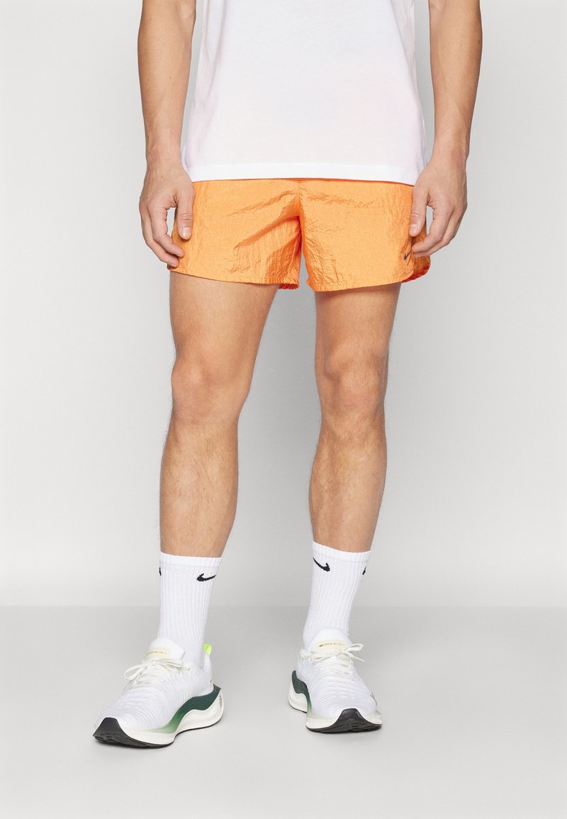 Homme debout portant un short de sport orange vif, des chaussettes blanches Nike, des chaussures de course blanches et un t-shirt blanc, sur un fond uni.