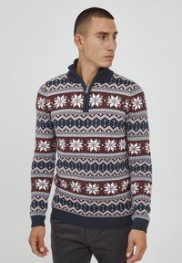 Pleteni pulover z maroon osnovo in belimi snežinkami ter geometrijskimi vzorci, dopolnjen z modrim zip ovratnikom in rebrastimi manšetami.