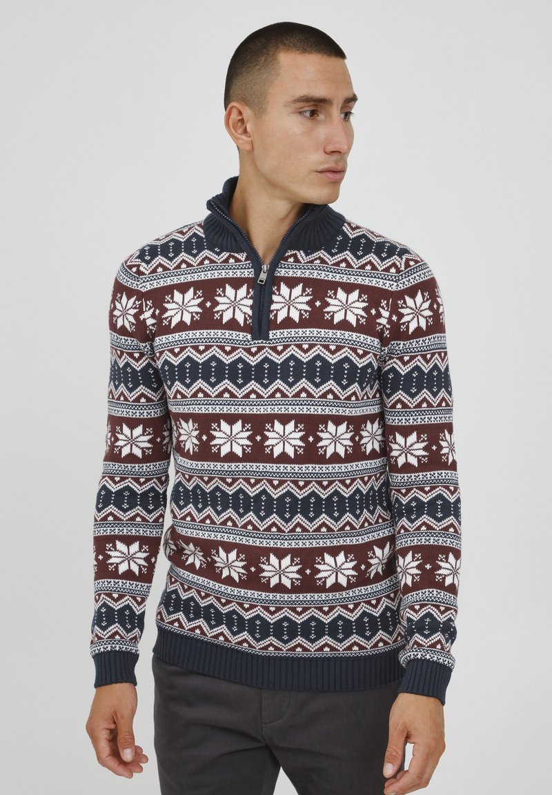 Pleteni pulover z maroon osnovo in belimi snežinkami ter geometrijskimi vzorci, dopolnjen z modrim zip ovratnikom in rebrastimi manšetami.