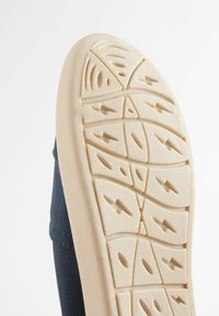 Semelle en caoutchouc avec des motifs texturés, de couleur beige, et de profonds rainures pour l'adhérence ; fixée à un dessus en toile bleu foncé.