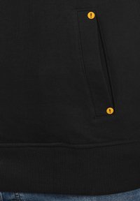 Sudadera negra con dobladillo acanalado, bolsillo lateral con acento circular amarillo y textura de tela suave. Diseño simple y funcional.
