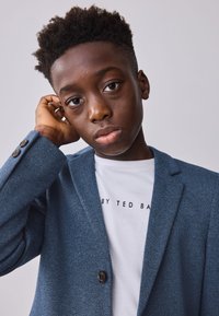 Blazer azul texturizado con botón negro, usado sobre una camiseta blanca con texto negro. El sujeto tiene el cabello corto y rizado y una expresión neutral.