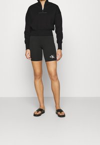 Sudadera negra con cremallera y logo bordado, combinada con pantalones cortos de ciclismo negros con un logo colorido. Llevada con chanclas negras.