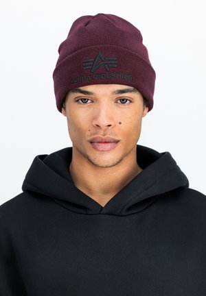 Maroon beanie hat with a ribbed texture and embroidered logo reading "ALPHA INDUSTRIES." Paired with a black hooded sweatshirt.