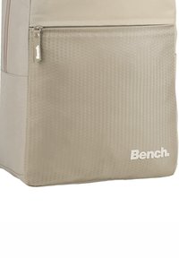 Beige backpack with a textured hexagonal pattern on the front, featuring a zip pocket and a white logo. Made of durable material.