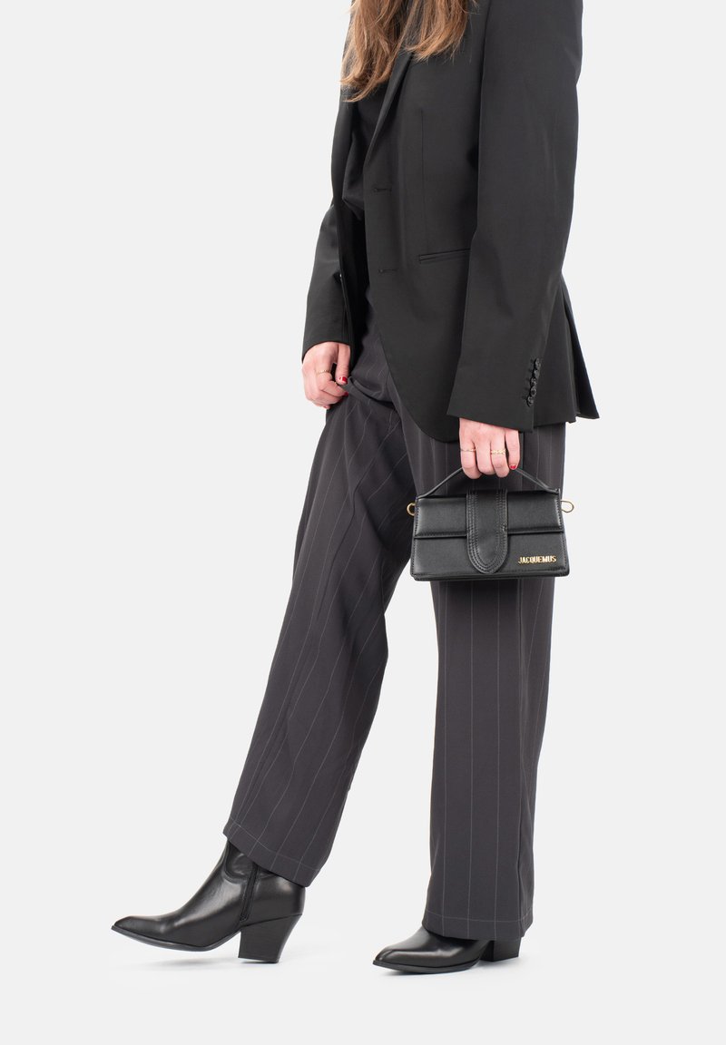 Sac en cuir noir au design structuré, avec un logo doré, tenu dans une main ; associé à un pantalon gris à rayures et des bottes noires à talons.