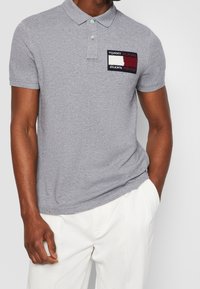 Mężczyzna ubrany w szary, krótki rękaw polo z naszywką logo "Tommy Hilfiger Shawn Mendes" i białe spodnie, z ręką w kieszeni.