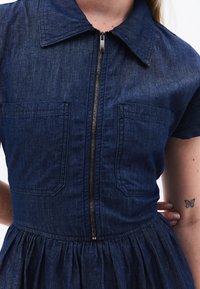 Denim-kjole med en tettsittende bodice, kortermede ermer, krage og en full, rynket skjørt; har to frontlommer og en glidelås foran.