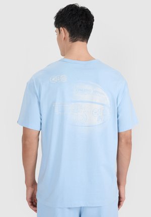 Mężczyzna ubrany w jasnoniebieski T-shirt z delikatną grafiką koszykówki i tekstem na plecach, stojący na tle jednolitego jasnego tła.