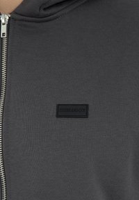 Sweatshirt zippé gris en tissu côtelé, avec un badge logo noir et une fermeture éclair argentée. Design simple avec une capuche et sans motifs supplémentaires.