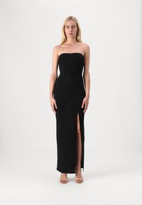 Črna brezramna obleka z oprijetim zgornjim delom in visokim razporkom na eni strani, izdelana iz teksturirane tkanine, v kombinaciji z nude petkami.