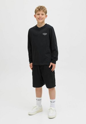 Zwart shirt met lange mouwen met logo, gecombineerd met zwarte cargoshorts, witte sneakers en witte sokken met zwarte letters.