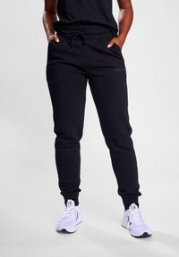 Schwarze Jogginghosen mit Kordelzugbund, elastischen Bündchen an den Knöcheln und Seitentaschen. Das Material wirkt weich mit einer glatten Textur.