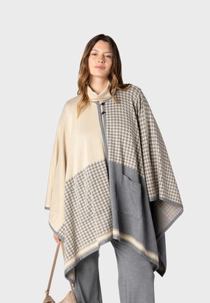 Femme portant un poncho beige et gris à motif pied-de-poule avec des boutons, tenant un sac à main beige, debout devant un fond uni.