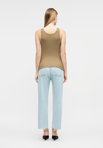 Γυναίκα με μακριά ξανθά μαλλιά, φορεί μπλούζα χωρίς μανίκια καφέ χρώματος, ανοιχτόχρωμα τζιν με φθαρμένες άκρες και μαύρα σανδάλια με τακούνι, στραμμένη προς τα πίσω.