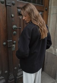 Mustat ylikokoiset bleiserit, joissa on jämäkkä muoto ja pehmeä rakenne, yhdistettynä vaaleisiin housuihin, puuoven taustalla.