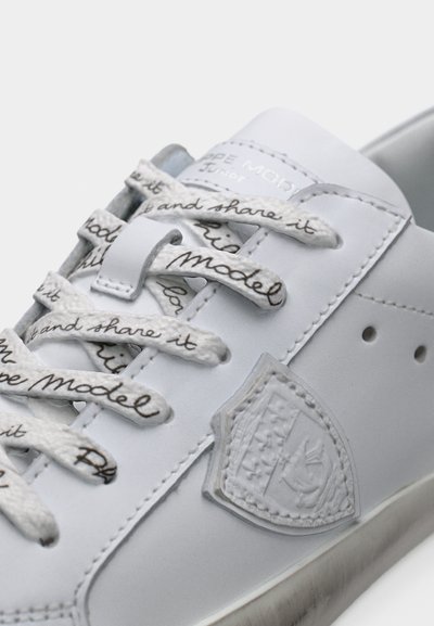 Λευκό δερμάτινο αθλητικό παπούτσι με κορδόνια τυπωμένα με γραφή, ανάγλυφη ετικέτα με λογότυπο και απαλό φινίρισμα. Σχεδίαση με χαμηλή κορυφή και στρογγυλεμένη μύτη.