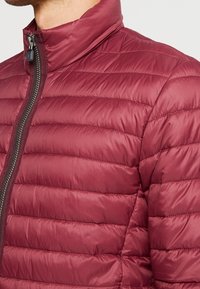 Bordeauxrote isolierte Jacke mit hohem Kragen, gesteppter Optik und vertikalen Streifen. Verfügt über einen durchgehenden Reißverschluss und eine glatte, glänzende Oberfläche.