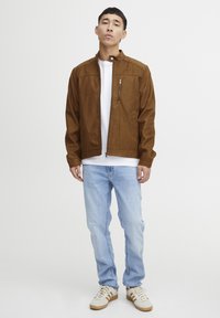 Veste en daim marron avec une poche zippée, associée à un t-shirt blanc, un jean bleu clair et des baskets crème avec des rayures marron.