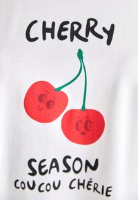 Weißes T-Shirt aus Stoff mit einem grafischen Print von zwei roten Kirschen mit Gesichtszügen, begleitet von dem Text "CHERRY SEASON coucou chérie" in Schwarz.